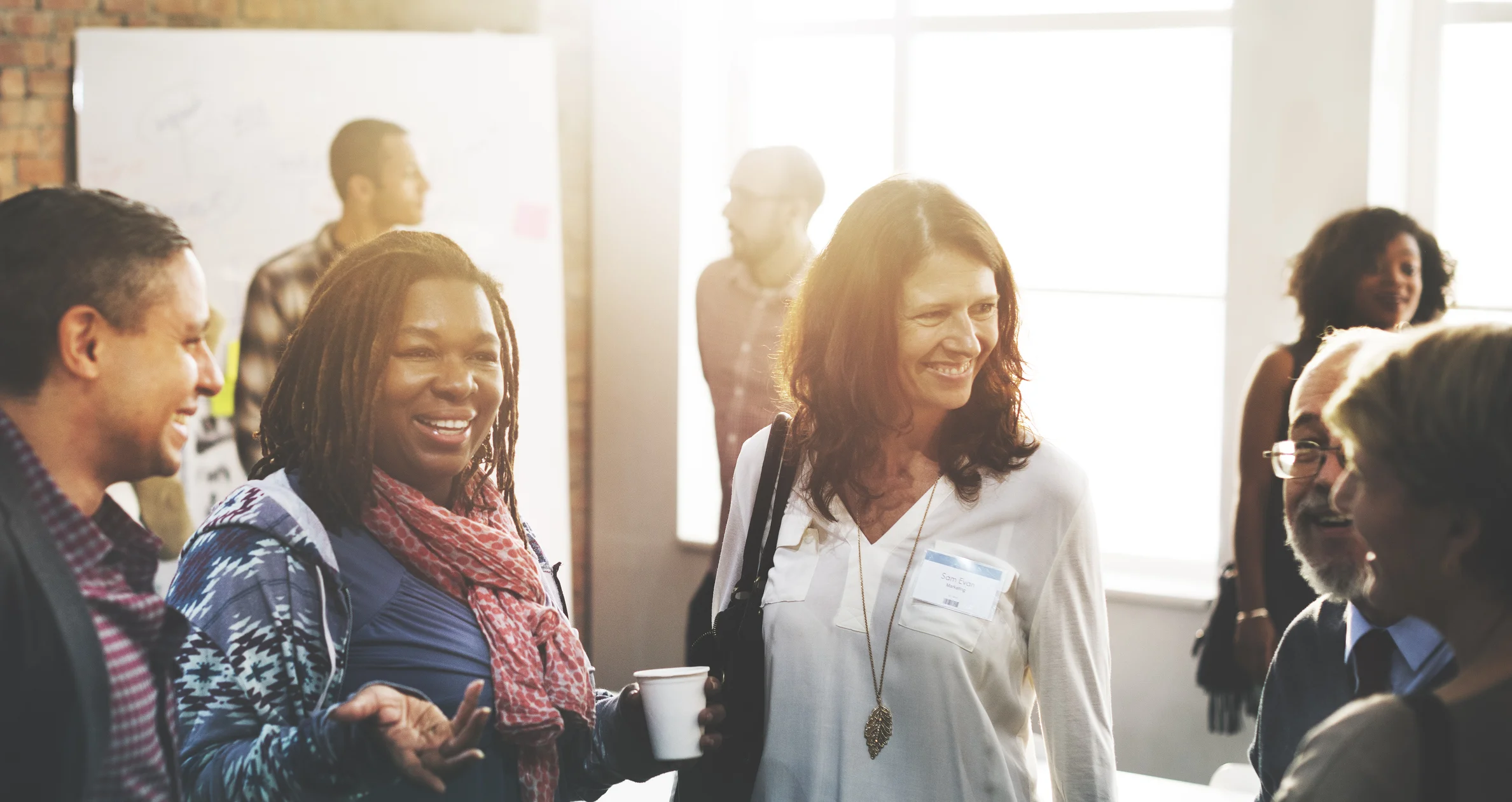 WELL SAID: TORONTO SPEECH THERAPY. A group of people, both men and women, are smiling and talking together in a bright room during a casual networking or social event. Some hold drinks, and sunlight streams through large windows in the background.