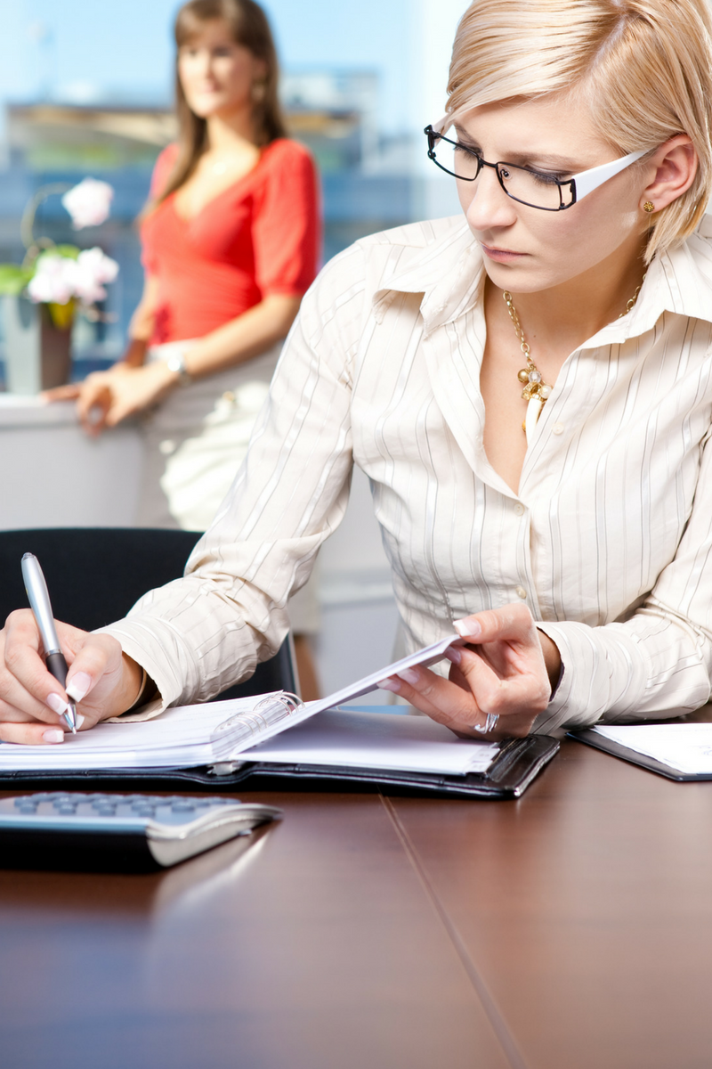 WELL SAID: TORONTO SPEECH THERAPY. A woman with glasses writes in a notebook at a desk, holding a document. Another woman stands in the background near a window, with sunlight in the office.