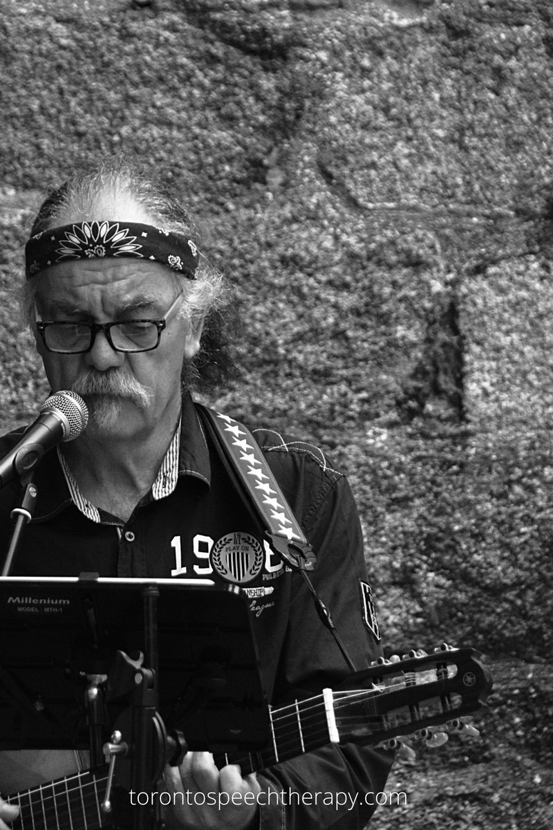 WELL SAID: TORONTO SPEECH THERAPY. A man with a mustache and glasses wearing a patterned headband plays guitar and sings into a microphone. He stands in front of a rough stone wall, with a music stand and guitar strap visible.
