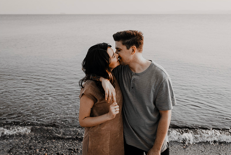 WELL SAID: TORONTO SPEECH THERAPY. A couple stands close together at the edge of a calm beach, smiling and touching noses. The woman has her arm around the mans shoulders, and the ocean extends into the distance behind them under a pale sky.
