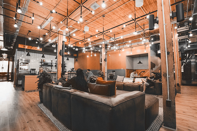 WELL SAID: TORONTO SPEECH THERAPY. A modern, open-concept lounge with leather sofas, wood floors, exposed ceilings, and hanging lights. Several people are sitting and talking, while others relax or work in the background. A kitchen area is visible.