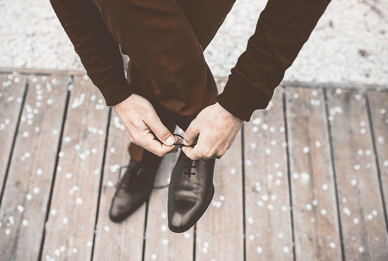 WELL SAID: TORONTO SPEECH THERAPY. A person wearing dark pants and a sweater bends down to tie the laces of a black dress shoe while standing on a wooden surface scattered with small white petals.