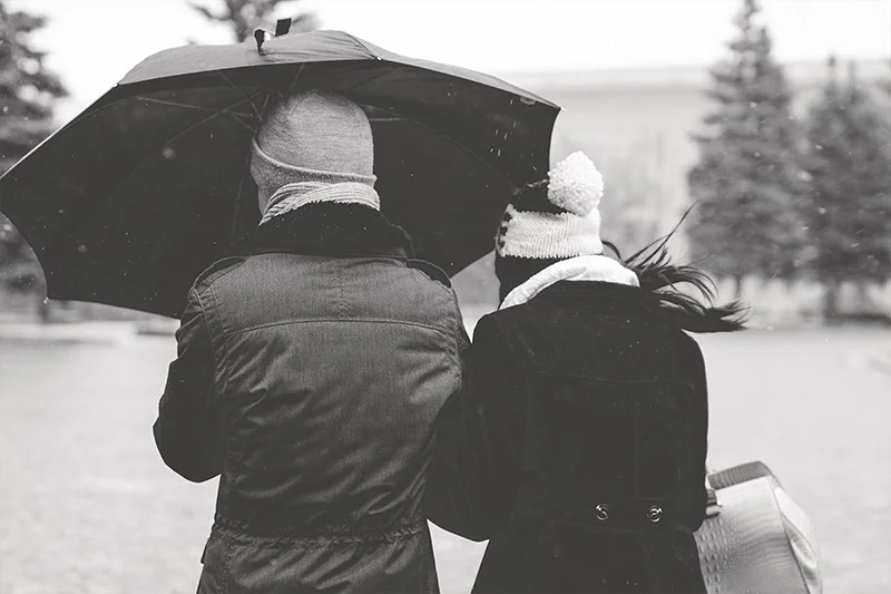 WELL SAID: TORONTO SPEECH THERAPY. Two people dressed in winter coats and hats walk side by side under a large umbrella on a snowy day, with trees in the background. The image is in black and white.