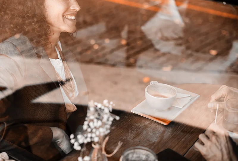 WELL SAID: TORONTO SPEECH THERAPY. A woman smiles while sitting at a table with a cup of coffee and flowers. The scene is viewed through a window, creating soft reflections and a warm, inviting atmosphere.