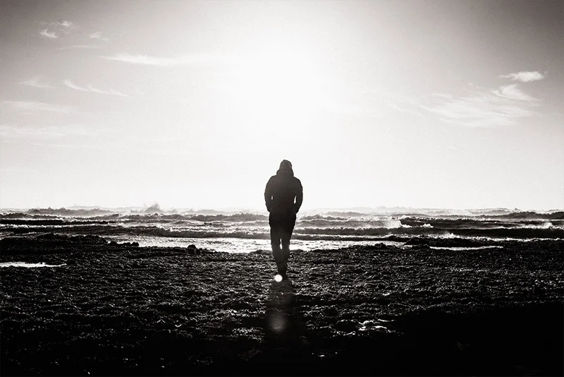 WELL SAID: TORONTO SPEECH THERAPY. A person stands alone on a rocky shore, facing the ocean with waves in the distance, silhouetted against a bright, hazy sky. The scene is in black and white, creating a moody, contemplative atmosphere.