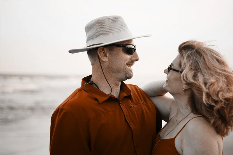 WELL SAID: TORONTO SPEECH THERAPY. A smiling couple wearing sunglasses and casual clothes stand close together on a beach, looking at each other affectionately. The man wears a wide-brimmed hat, and the woman has her hand on his shoulder.