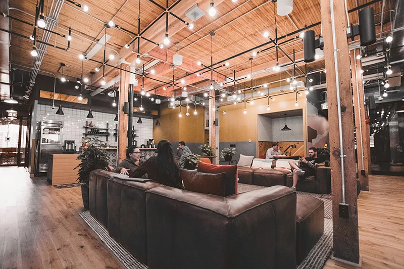 WELL SAID: TORONTO SPEECH THERAPY. Modern coworking space with exposed wooden beams, hanging lights, indoor plants, and people sitting and talking on brown leather couches; open kitchen area in the background.