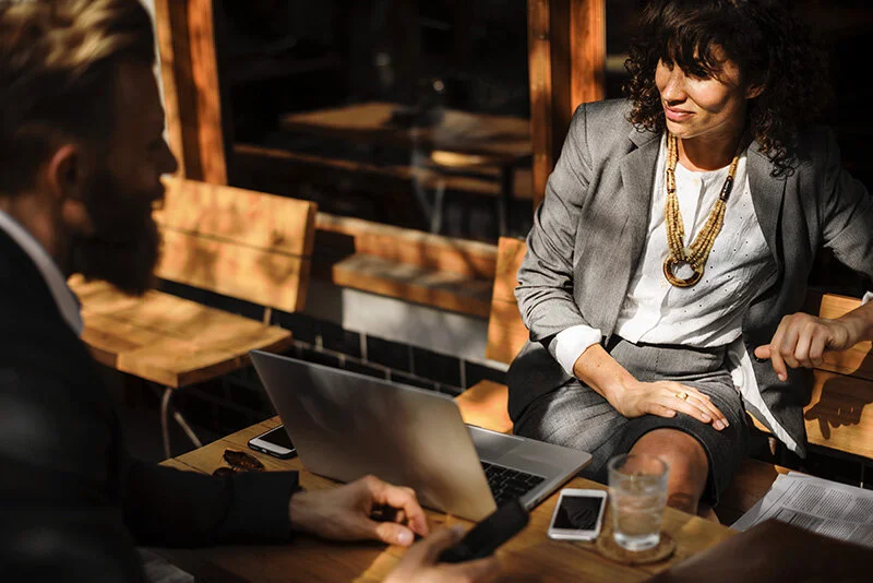 WELL SAID: TORONTO SPEECH THERAPY. Two people in business attire sit at an outdoor café table with a laptop, smartphones, and drinks, engaged in conversation. Sunlight casts shadows across the scene.