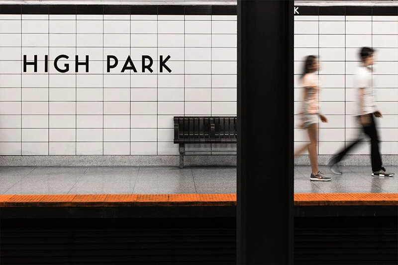 WELL SAID: TORONTO SPEECH THERAPY. Two people walk past each other on a subway platform in front of a white tiled wall with HIGH PARK written on it. A black bench is positioned against the wall.