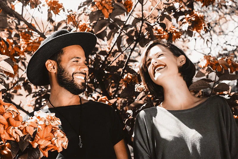 WELL SAID: TORONTO SPEECH THERAPY. A man wearing a black hat and a woman with short hair are standing outdoors, smiling and laughing together, surrounded by orange and brown foliage.