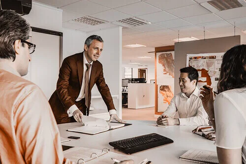 WELL SAID: TORONTO SPEECH THERAPY. A man in a suit stands at a conference table, presenting to three colleagues who are seated and listening in a modern office setting. Papers, a keyboard, and notebooks are on the table.