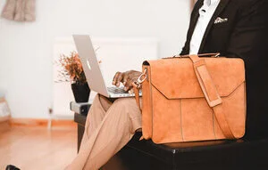 WELL SAID: TORONTO SPEECH THERAPY. A person in business attire sits on a black bench, typing on a laptop, with a tan leather briefcase resting on their lap. The background shows a light-colored room with a potted plant.