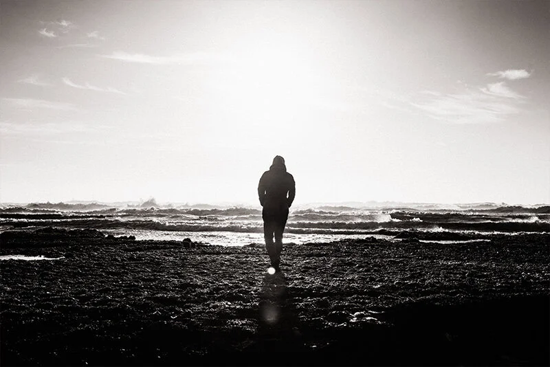 WELL SAID: TORONTO SPEECH THERAPY. A person in a hooded jacket walks alone on a rocky shore toward the ocean, silhouetted against a bright, hazy sky as sunlight reflects off the water.