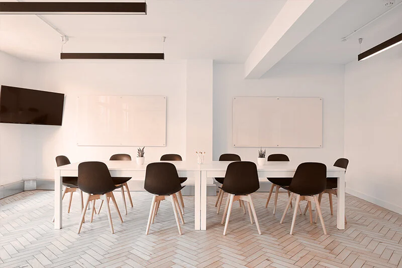 WELL SAID: TORONTO SPEECH THERAPY. A modern conference room with a long white table, ten black chairs, two whiteboards on the wall, a wall-mounted TV, and light wood herringbone flooring. The room is bright and minimalistic.