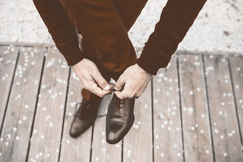 WELL SAID: TORONTO SPEECH THERAPY. A person wearing brown pants is seen from above, bending down to tie the laces of a black dress shoe while standing on a wooden deck.