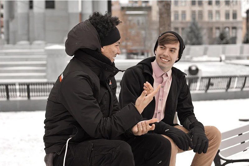 WELL SAID: TORONTO SPEECH THERAPY. Two young men wearing winter jackets and gloves sit on a snowy bench outdoors, engaged in a lively, friendly conversation. One gestures with his hands while the other smiles, listening attentively.