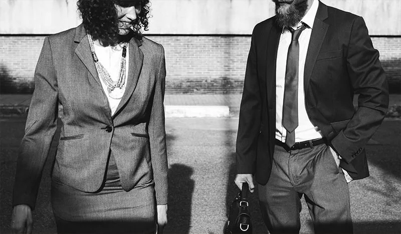 WELL SAID: TORONTO SPEECH THERAPY. A woman and a man in business attire walk outside. The woman wears a skirt suit and necklace, and the man wears a suit and tie while holding a bag. Both appear to be engaged in conversation. The photo is in black and white.