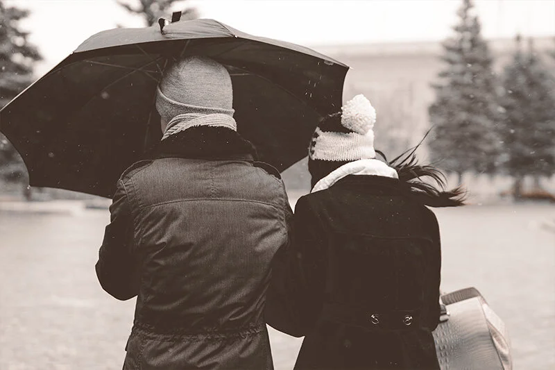 WELL SAID: TORONTO SPEECH THERAPY. Two people in winter coats and hats walk closely together under an umbrella on a snowy day, with evergreen trees in the background. The image is in black and white.