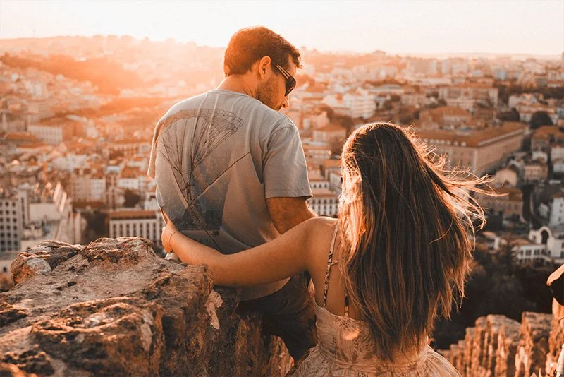 WELL SAID: TORONTO SPEECH THERAPY. A couple sits on a rocky ledge overlooking a sunlit cityscape at sunset. The woman has her arm around the mans back as they enjoy the scenic view together.
