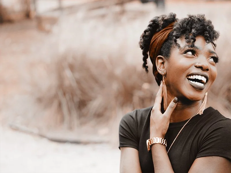 WELL SAID: TORONTO SPEECH THERAPY. A young woman with curly hair and a headband smiles brightly while looking to the side. She wears a dark shirt, gold earrings, a watch, and rests one hand on her neck. The background is blurred and natural.