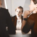 WELL SAID: TORONTO SPEECH THERAPY. Three women sitting at a table, two in the foreground with blurred faces, and one facing the camera with a serious expression, suggesting a meeting or job interview setting.