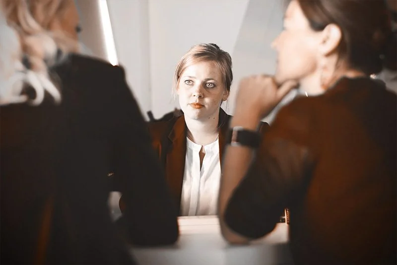 WELL SAID: TORONTO SPEECH THERAPY. Three women sitting at a table, two in the foreground with blurred faces, and one facing the camera with a serious expression, suggesting a meeting or job interview setting.