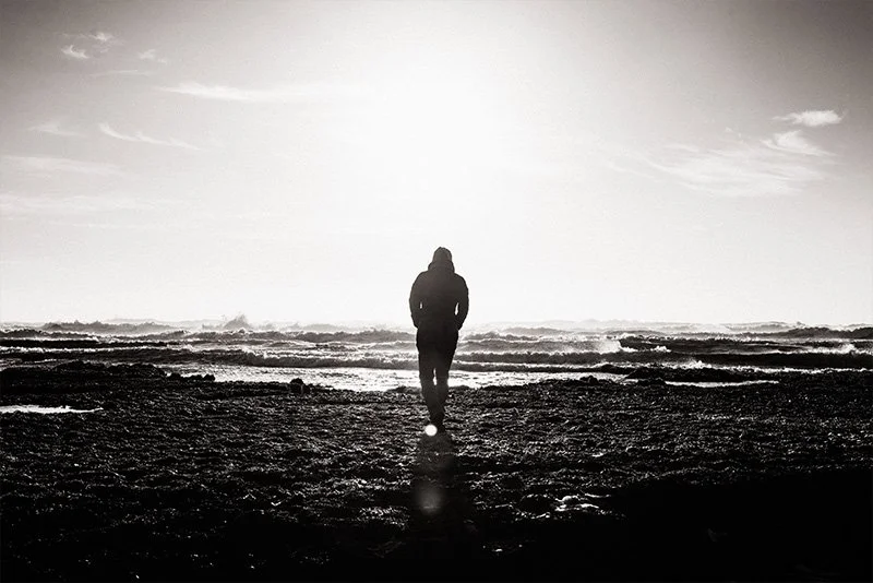 WELL SAID: TORONTO SPEECH THERAPY. A person in a hooded jacket walks alone on a rocky shore toward the ocean, with bright sunlight creating a dramatic silhouette and shimmering reflections on the water.
