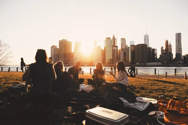 WELL SAID: TORONTO SPEECH THERAPY. A group of people have a picnic on grass at sunset, with food and blankets, overlooking a river and a city skyline with tall buildings in the background.
