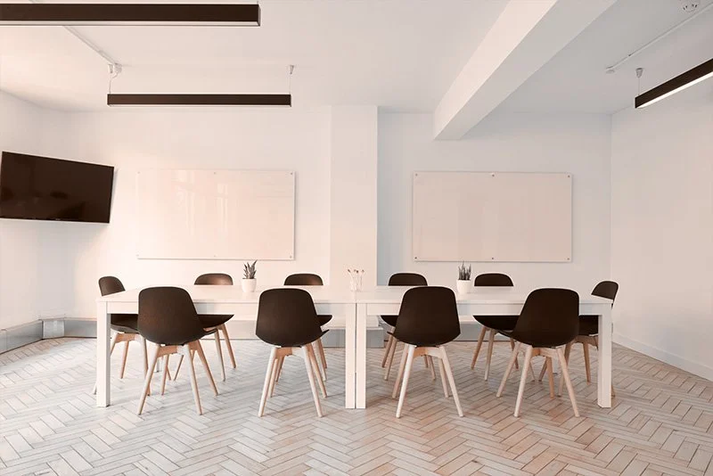WELL SAID: TORONTO SPEECH THERAPY. A modern, minimalist conference room with two white tables, eight black chairs, two whiteboards on the wall, a flat-screen TV, and small potted plants on the tables. The room has light-colored walls and herringbone flooring.