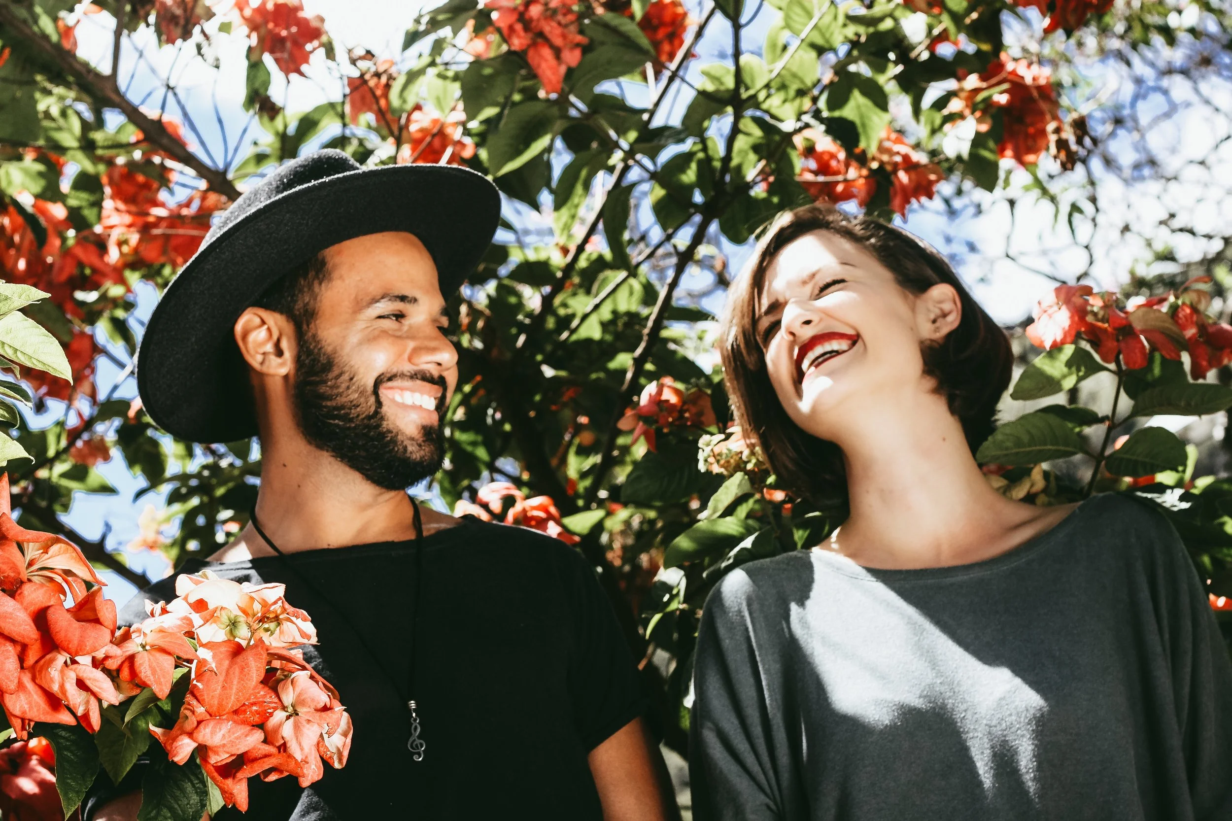 WELL SAID: TORONTO SPEECH THERAPY. A man with a beard and hat and a woman with short hair are smiling and laughing together outdoors, surrounded by lush greenery and red flowers under a bright sky.