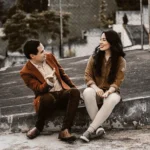 WELL SAID: TORONTO SPEECH THERAPY. Two people sit on a rooftop, facing each other and smiling. The man gestures as he talks, while the woman listens, both appearing relaxed and engaged in conversation. A chain-link fence and trees are visible in the background.