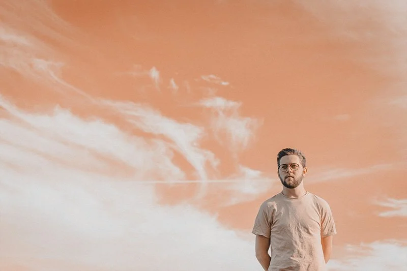 WELL SAID: TORONTO SPEECH THERAPY. A man with glasses and a beard stands outdoors against a sky with wispy clouds and an orange tint. He is wearing a light-colored t-shirt and looks slightly to the side.