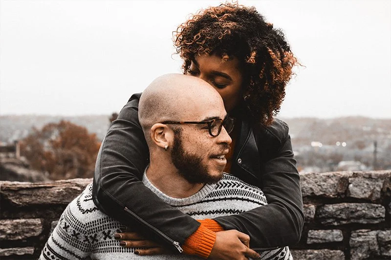 WELL SAID: TORONTO SPEECH THERAPY. A woman hugs a man from behind and kisses his head. The man, wearing glasses and a patterned sweater, smiles. They stand in front of a stone wall with a blurred outdoor background.