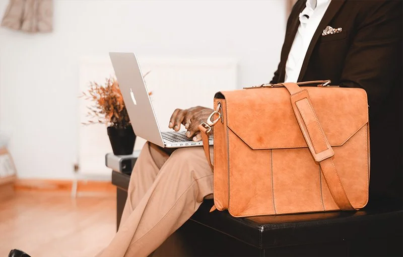 WELL SAID: TORONTO SPEECH THERAPY. A person in business attire sits with a laptop on their lap, typing. A tan leather briefcase rests beside them on the bench. The background is an indoor setting with neutral colors.