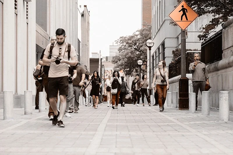 WELL SAID: TORONTO SPEECH THERAPY. A group of people walk along a city sidewalk, some looking at their phones or cameras. Buildings line both sides, and a pedestrian crossing sign is visible on the right. The scene appears casual and urban.