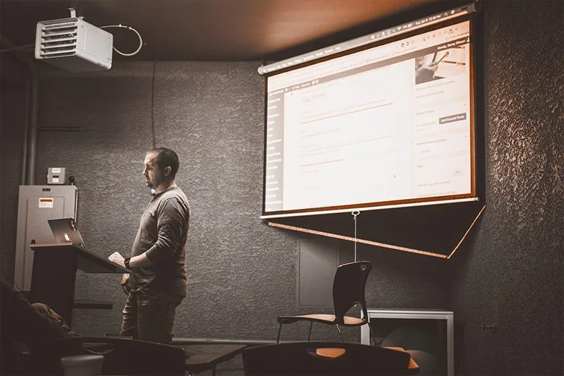 WELL SAID: TORONTO SPEECH THERAPY. A man stands at a podium with a laptop, giving a presentation in a dimly lit room. A large screen behind him displays a website interface with text and images. Empty chairs are visible in the foreground.