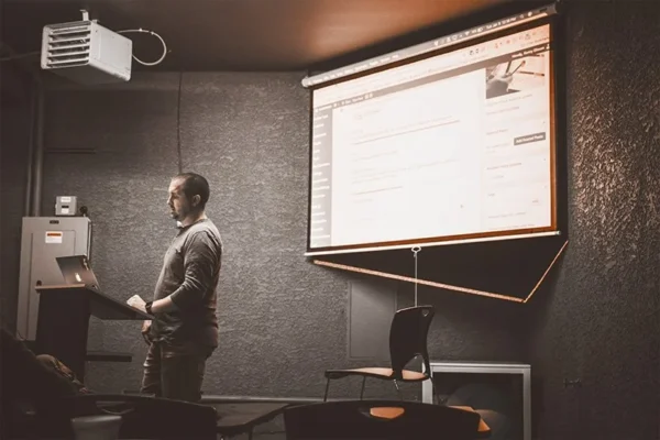WELL SAID: TORONTO SPEECH THERAPY. A man stands at a podium with a laptop, giving a presentation on speech-language pathology in a dimly lit room. A large screen behind him displays a website or document, with empty chairs in front of him.
