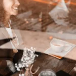 WELL SAID: TORONTO SPEECH THERAPY. A woman smiling at a café table in Ontario, seen through a window, with a cup of coffee and a small vase of flowers on the wooden table. Another person’s hands are visible, suggesting a warm meeting for adults with ASD.