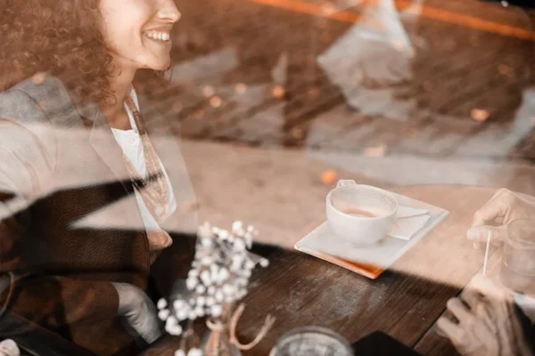 WELL SAID: TORONTO SPEECH THERAPY. A woman smiling at a café table in Ontario, seen through a window, with a cup of coffee and a small vase of flowers on the wooden table. Another person’s hands are visible, suggesting a warm meeting for adults with ASD.
