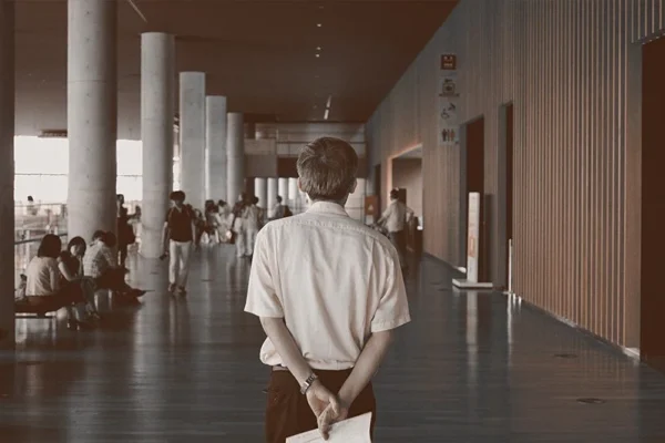 WELL SAID: TORONTO SPEECH THERAPY. A man stands with his back to the camera, holding papers behind him in a spacious, modern hallway in Ontario, with people sitting and walking in the background. The scene has a calm, slightly vintage tone.