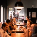 WELL SAID: TORONTO SPEECH THERAPY. People sit at long tables in a warmly lit Toronto restaurant, engaged in conversation and enjoying drinks, with large windows on one side and hanging lights above.
