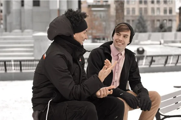 WELL SAID: TORONTO SPEECH THERAPY. Two young men in winter clothing sit on a bench in snowy Ontario, engaged in a lively conversation and smiling at each other. Buildings and trees can be seen in the background.