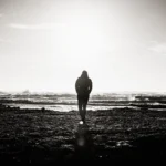 WELL SAID: TORONTO SPEECH THERAPY. A person stands alone on a rocky shore in Ontario, facing the ocean with waves in the distance. The image is in black and white, and the sun above the horizon creates a dramatic silhouette.