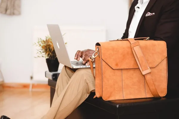 WELL SAID: TORONTO SPEECH THERAPY. A person in a suit sits on a bench using a laptop, with a tan leather briefcase beside them. The indoor setting, decorated with a small plant, could be an Ontario workspace designed for adults with ADHD seeking focus and productivity.