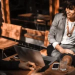 WELL SAID: TORONTO SPEECH THERAPY. Two people in business attire sit at an outdoor table with a laptop and smartphones, having a conversation in sunlight. The woman, facing the camera, discusses ASD support for adults in Ontario while the man remains mostly out of focus.