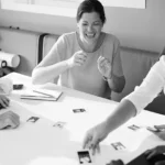 WELL SAID: TORONTO SPEECH THERAPY. Four people sit around a table in Toronto, smiling and interacting with small photos spread out in front of them. One woman laughs while another reaches toward the center. Notebooks and a coffee mug are also on the table.