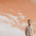 WELL SAID: TORONTO SPEECH THERAPY. A man in a light-colored t-shirt stands outdoors against a dramatic sky with orange and white clouds in Toronto, looking slightly to the side with a neutral expression.