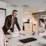WELL SAID: TORONTO SPEECH THERAPY. A man in a suit stands and speaks to three colleagues seated around a conference table in a modern office, discussing strategies and challenges for adults with ADHD; documents, a keyboard, and notebooks are visible on the table.
