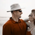 WELL SAID: TORONTO SPEECH THERAPY. A man in a sunhat and sunglasses smiles at a woman with wavy hair and glasses at the beach near Toronto. They are facing each other, both in orange clothing, with the ocean in the background.