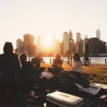 WELL SAID: TORONTO SPEECH THERAPY. A group of adults have a picnic on a grassy area near the water at sunset, with Toronto’s city skyline and tall buildings in the background. The sun is low, casting a warm glow over the peaceful scene.
